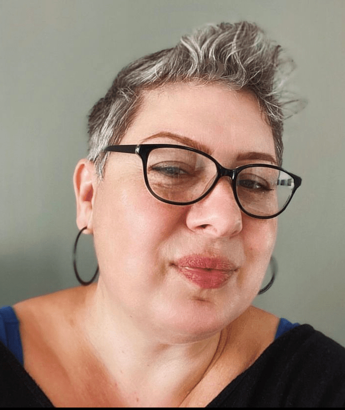 Photo of a white woman with short grey hair, black framed glasses and black hoop earrings